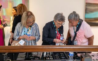 Three women closely examining jewellery pieces at the display table at Baycrest’s art event.