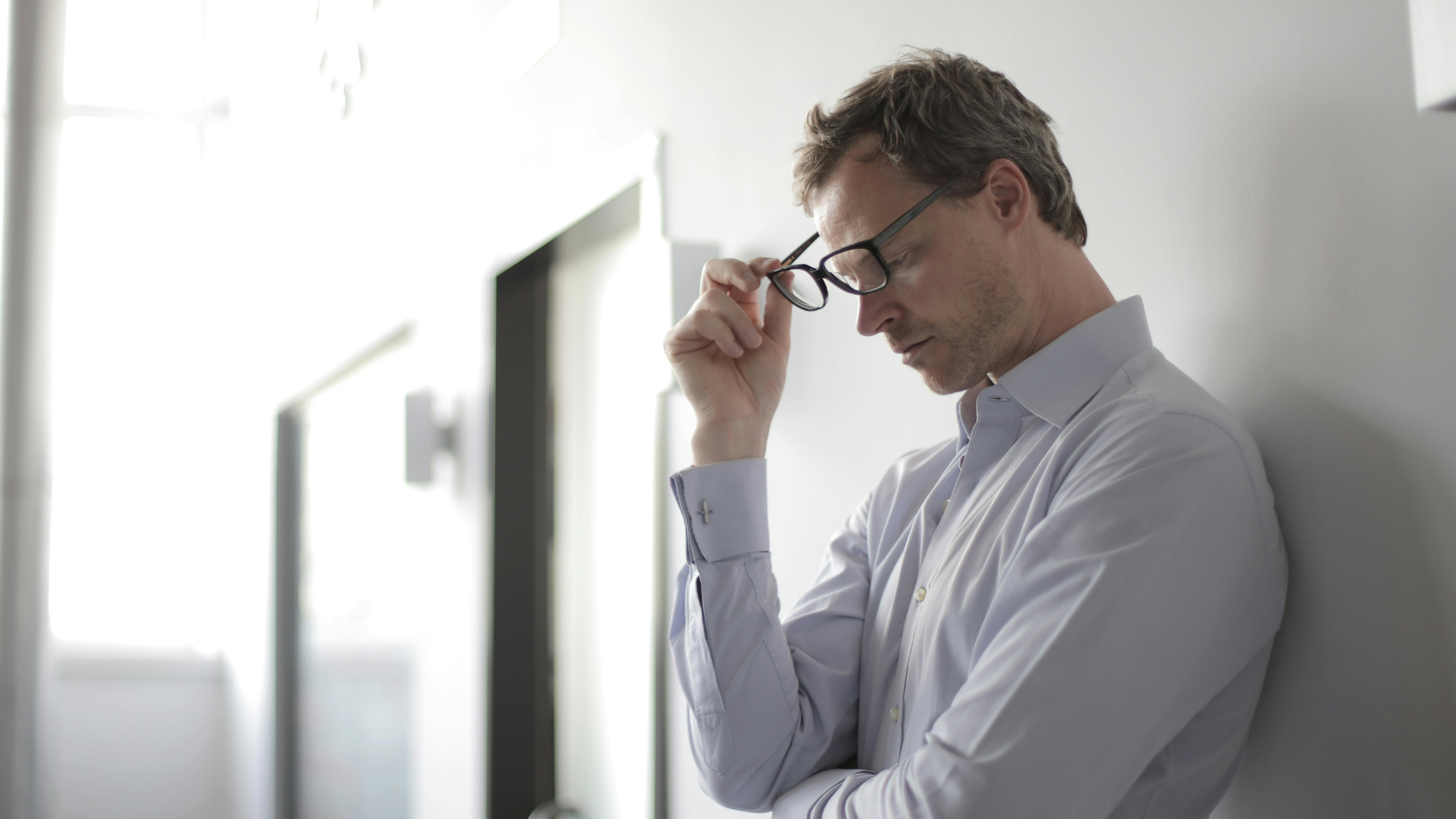 Man leaning against wall removing glasses looking frustrated